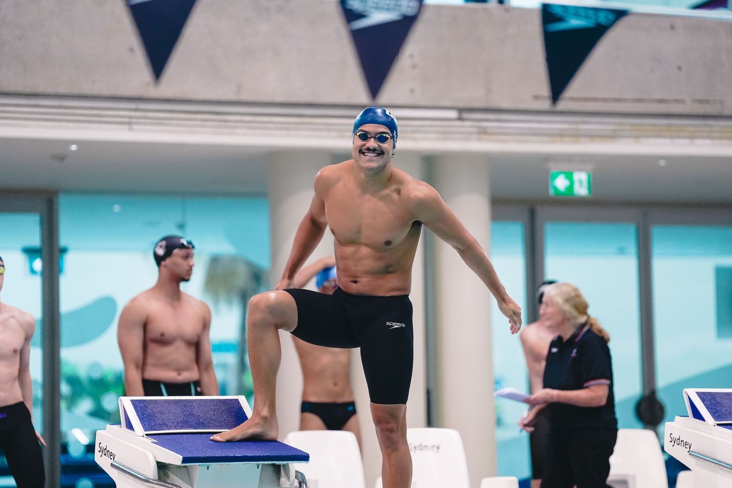 Swimmers on starting blocks prepare to dive into a pool during a competitive swimming event in an indoor arena.
