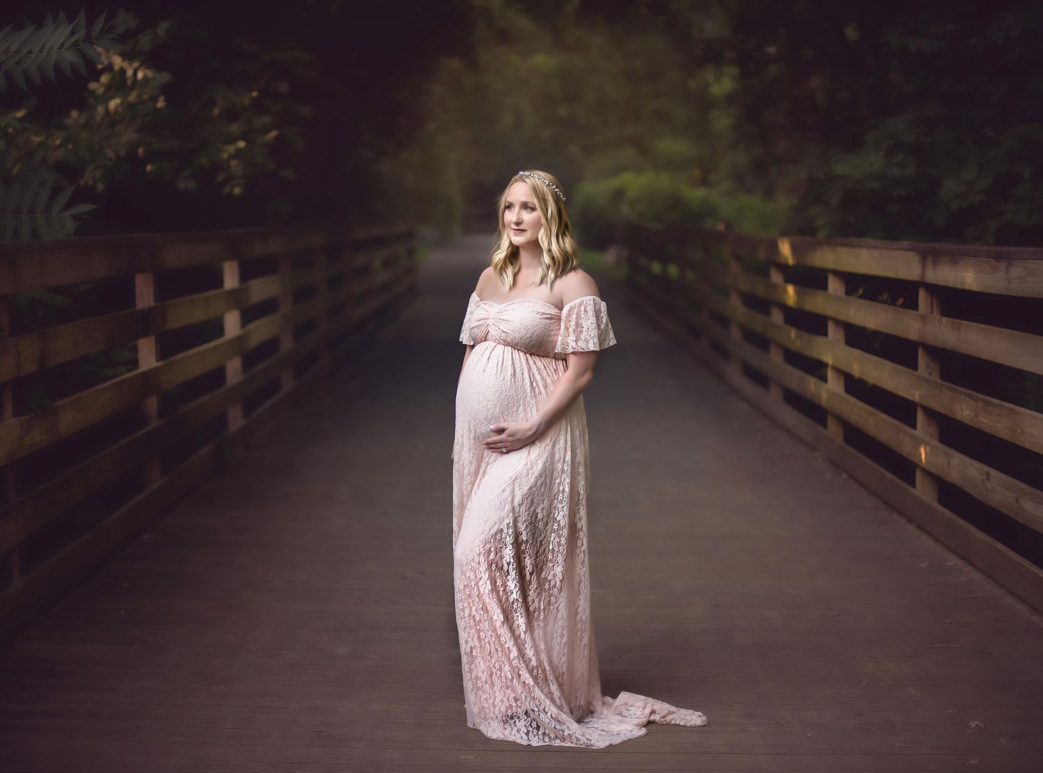 A maternity photo on a wooden bridge with a woman in a blush lace gown.