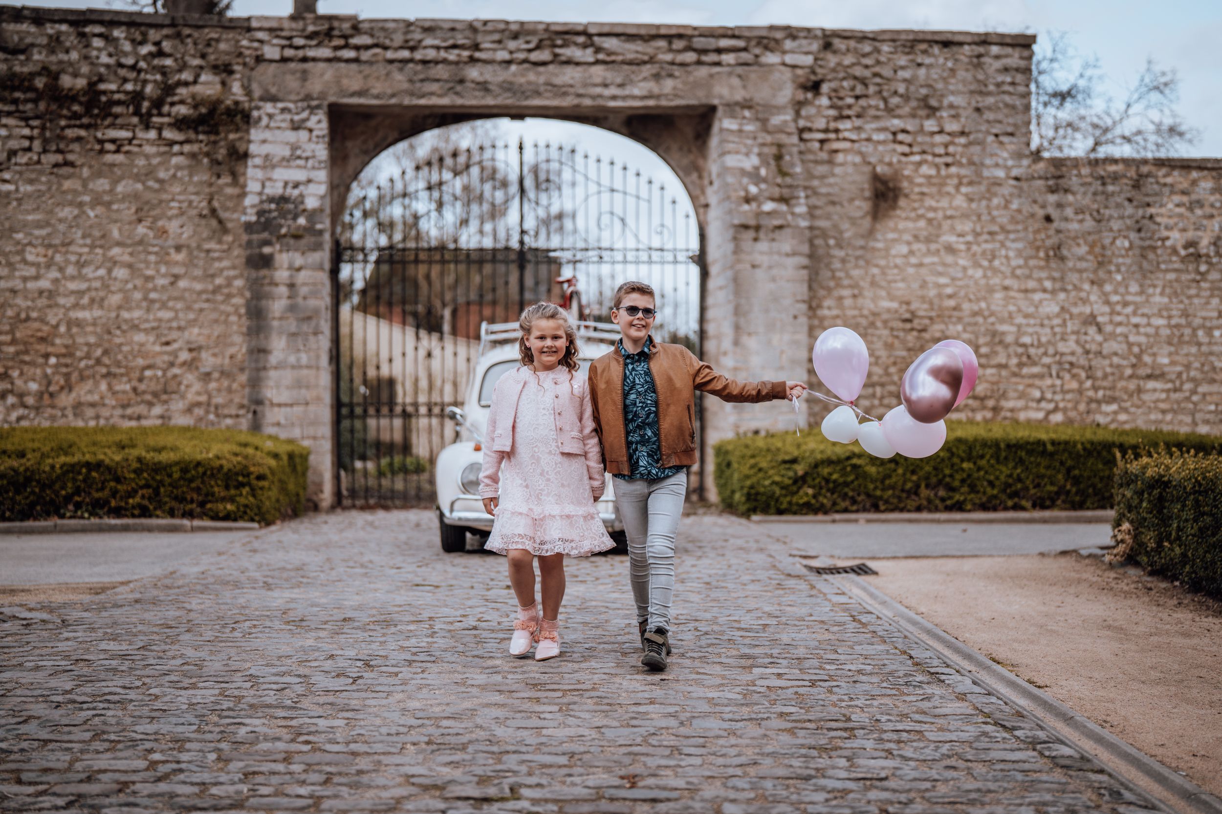 Een stel loopt hand in hand over een stenen pad bij een oude bakstenen boog met roze ballonnen die achter hen zweven.