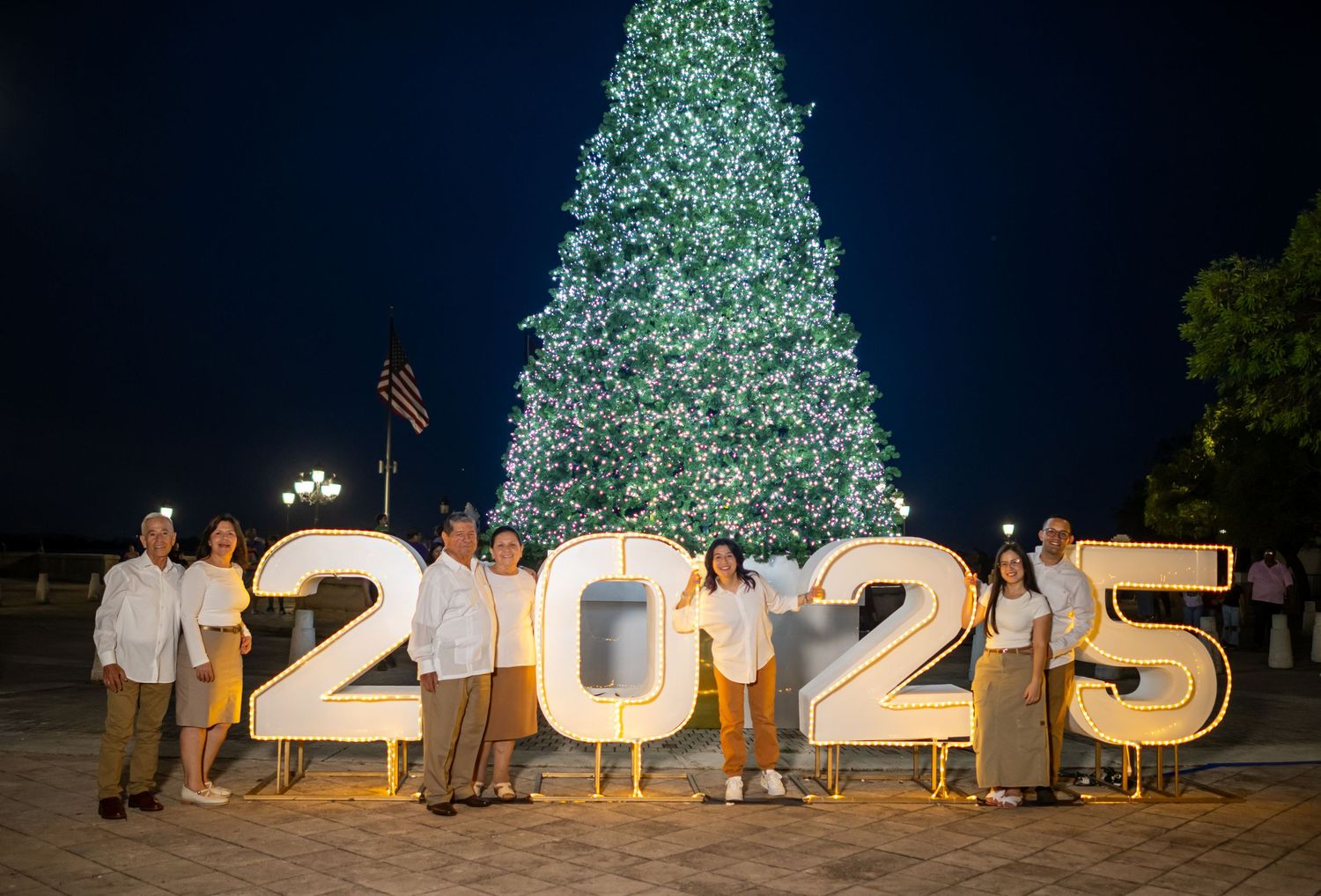 A family poses with illuminated 2025 numbers in front of a large lit Christmas tree at night.