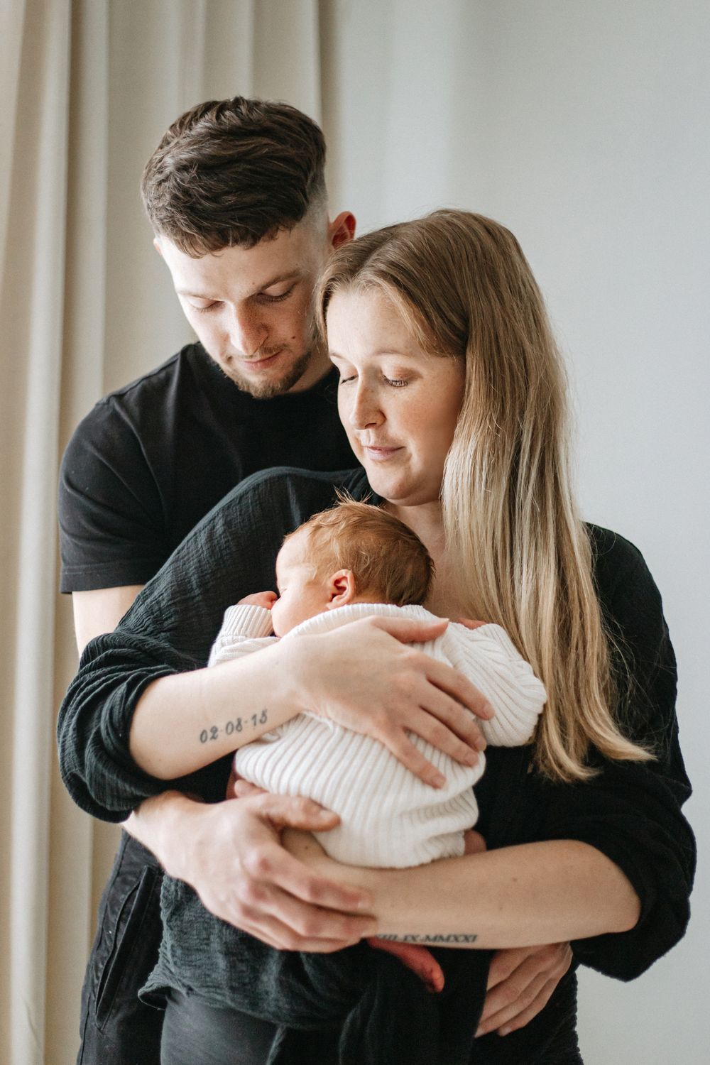 A couple in black sweaters tenderly hold their newborn baby wrapped in white against a light curtained background.