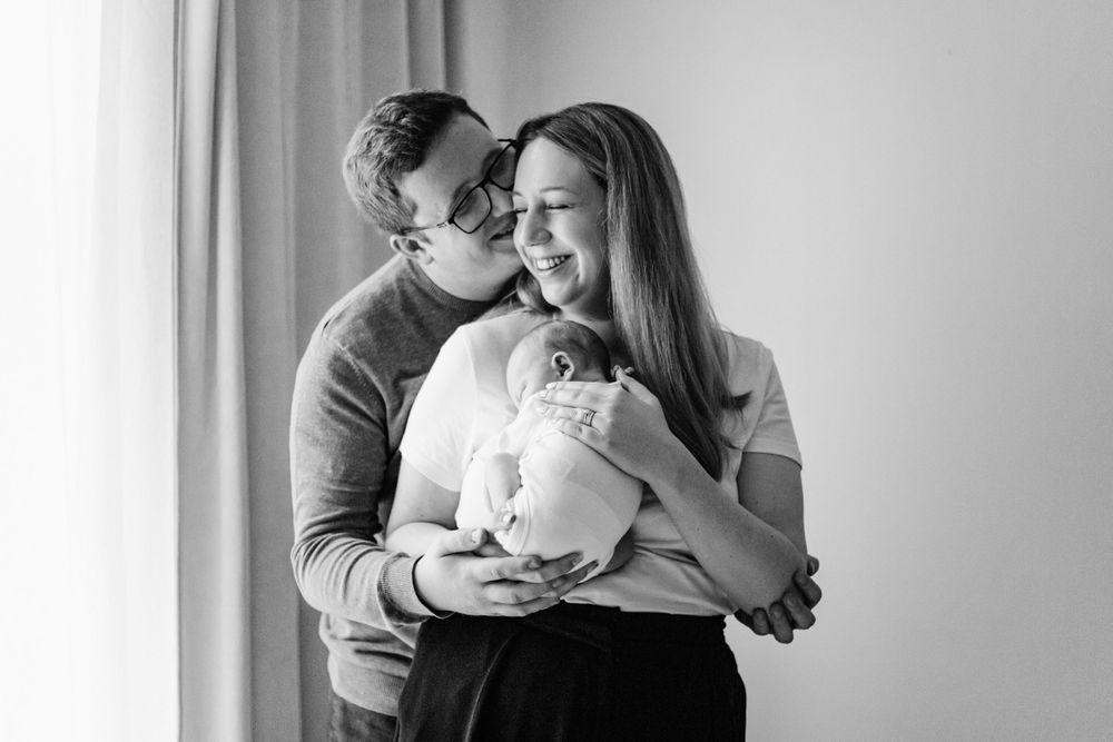 A loving couple embraces while holding their newborn baby in a tender black and white portrait by a window.