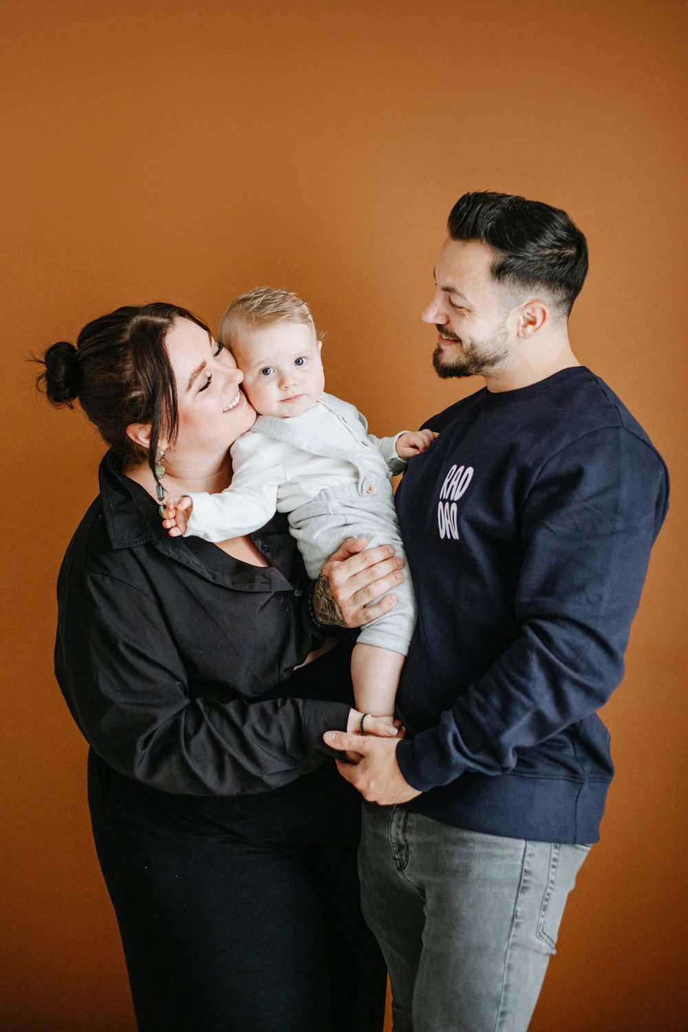 Family portrait of parents in black sweaters holding their baby in white against a warm brown studio backdrop.
