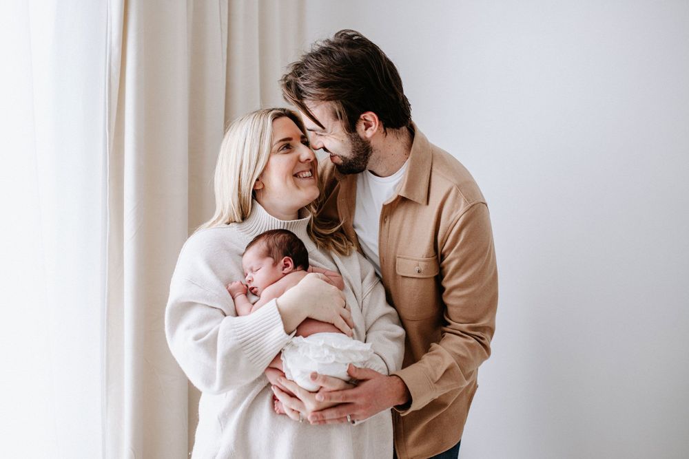A couple in cosy sweaters sharing an intimate moment while holding their sleeping newborn baby by a curtained window.