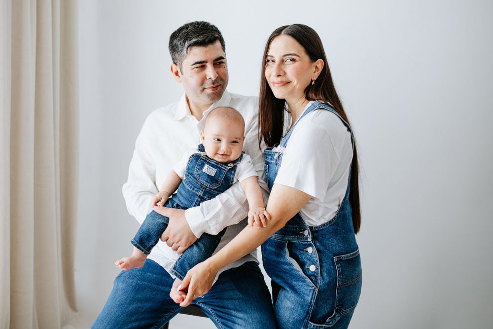 A family wearing matching denim overalls sits together against a white wall.