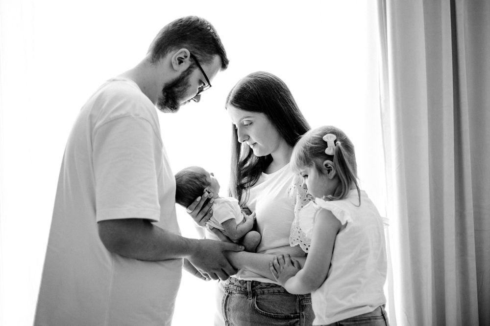 A black and white family portrait shows parents with their baby and young daughter by a bright window.