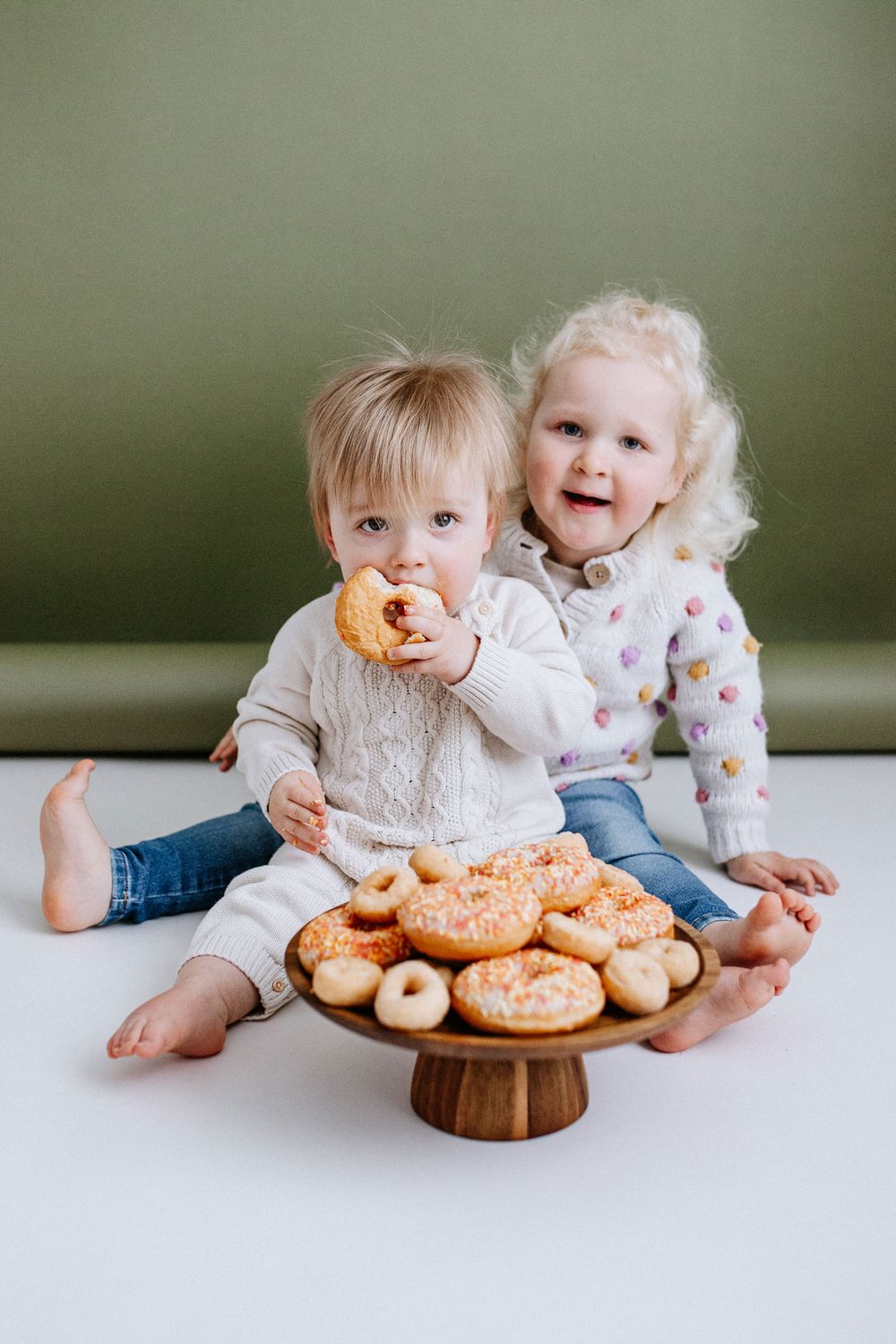 Two small children sit on floor with wooden cake stand full of glazed donuts as one takes a happy bite.