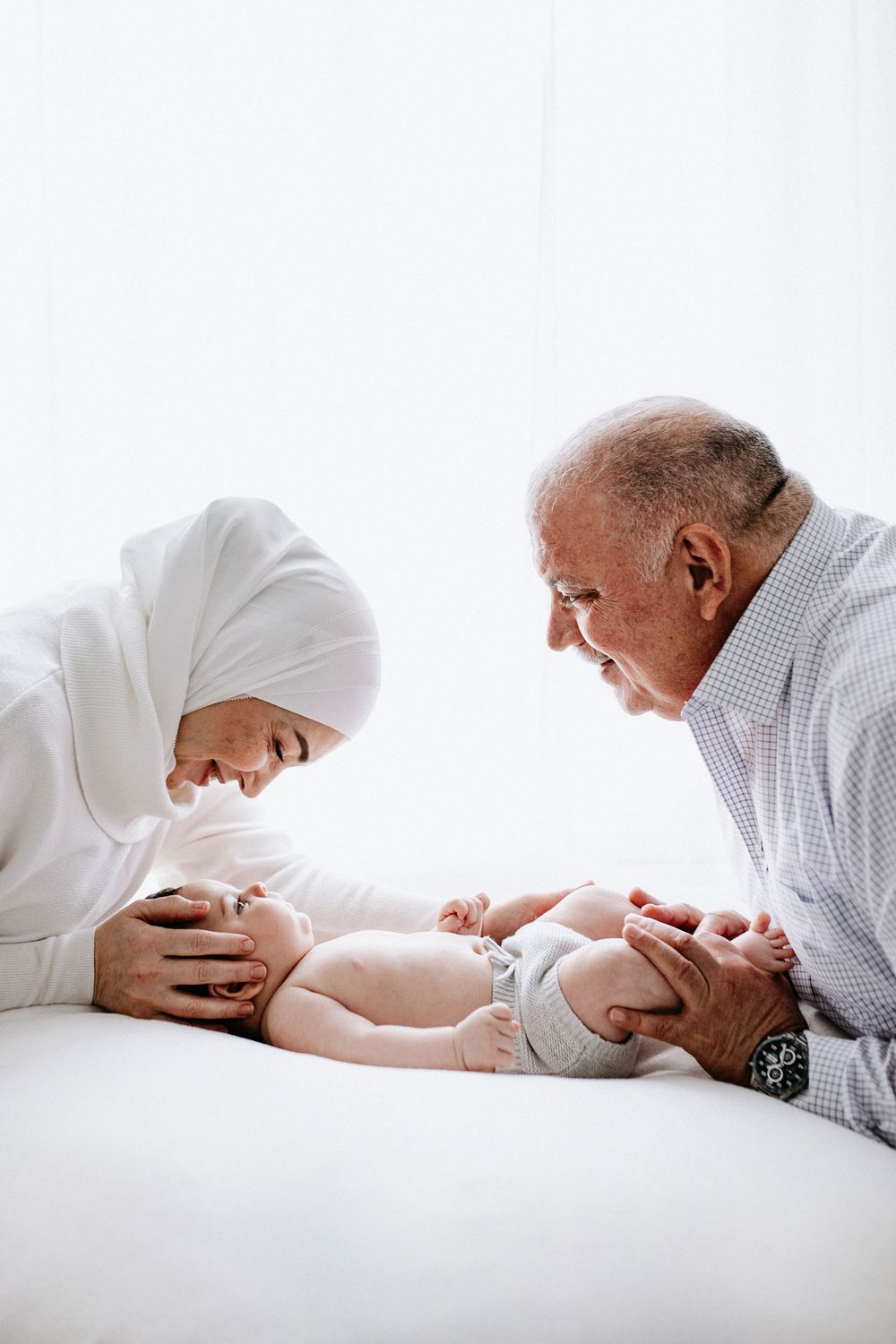 Grandparents lovingly interact with their infant grandchild while lying on a white surface together.
