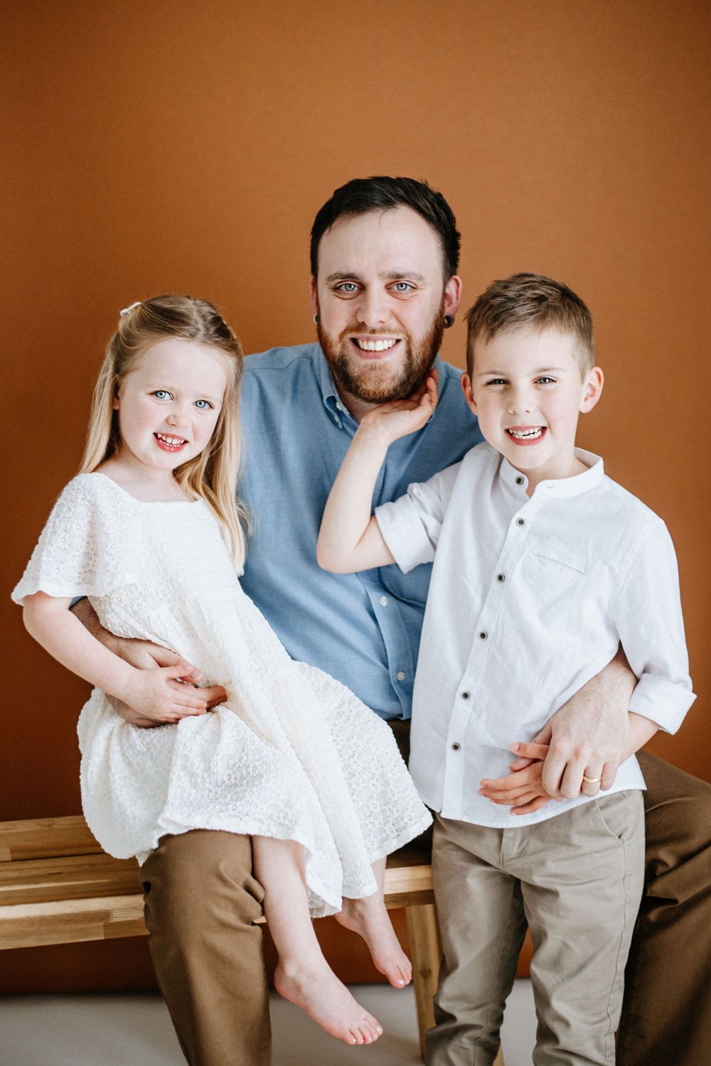 A family portrait against a brown background with coordinated white and neutral outfits sitting close together.