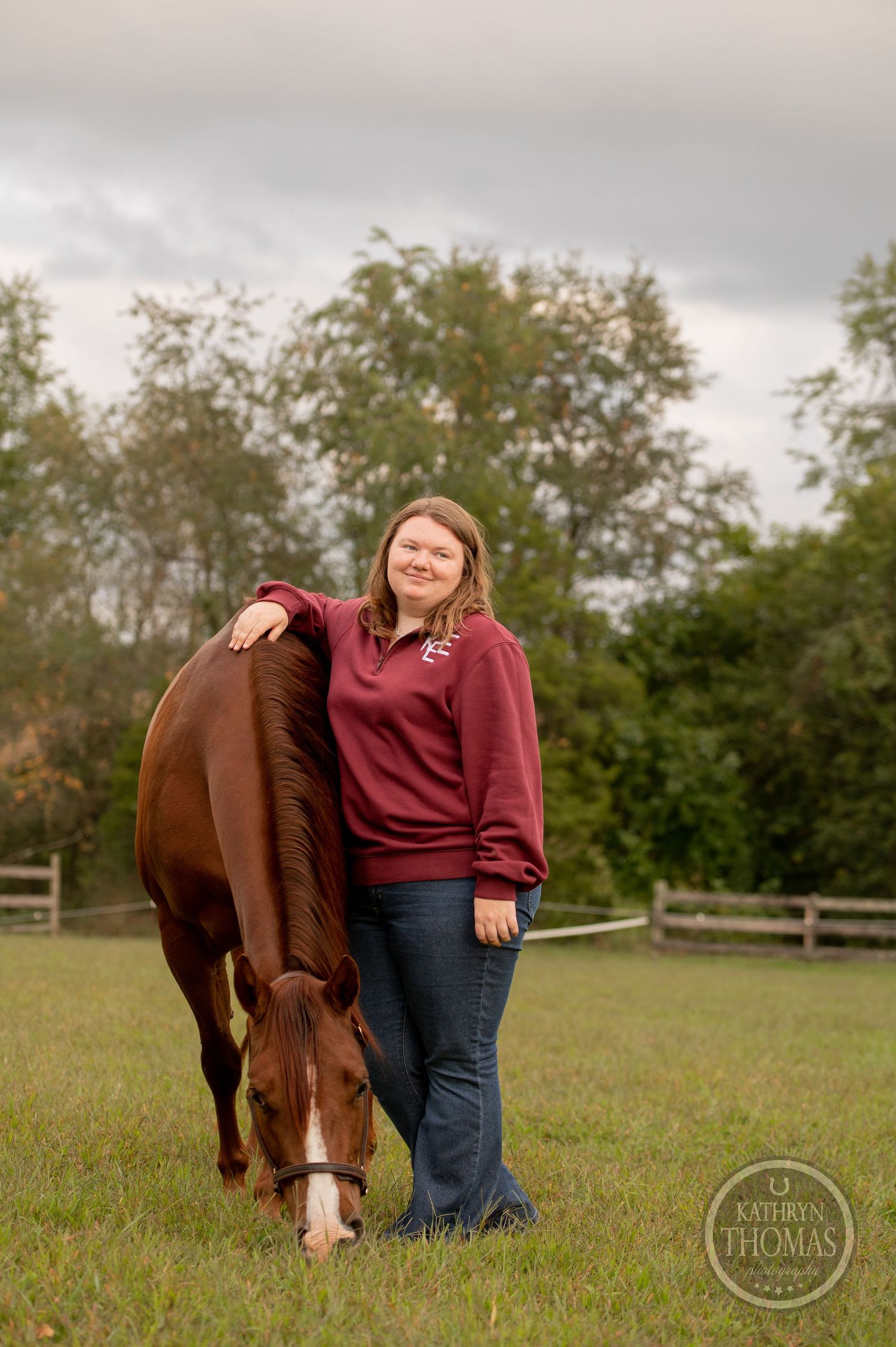 A chestnut horse grazes while someone in a burgundy sweater stands nearby in a pastoral setting.