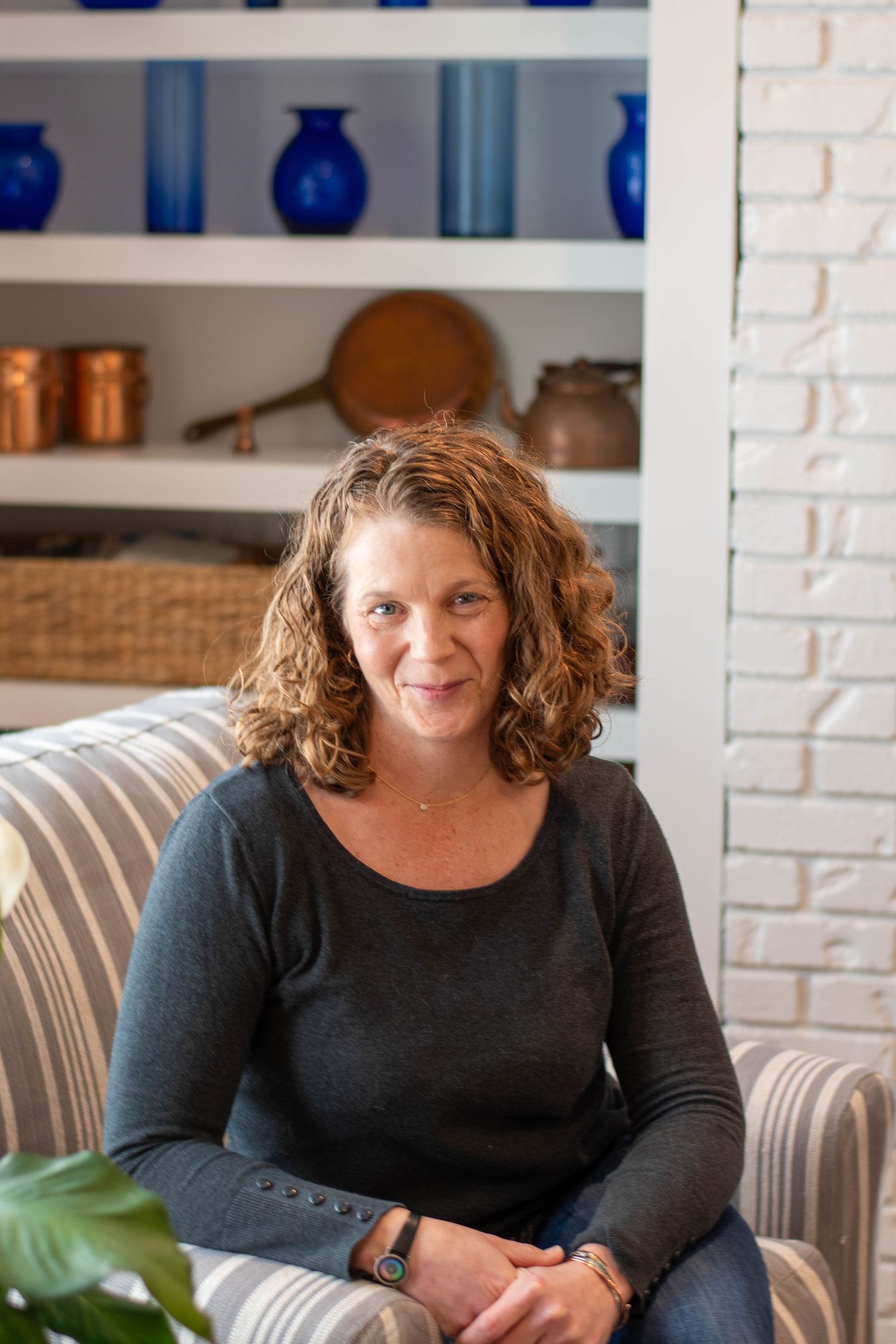 A woman in a black sweater sitting casually on a couch with blue decor in the background.