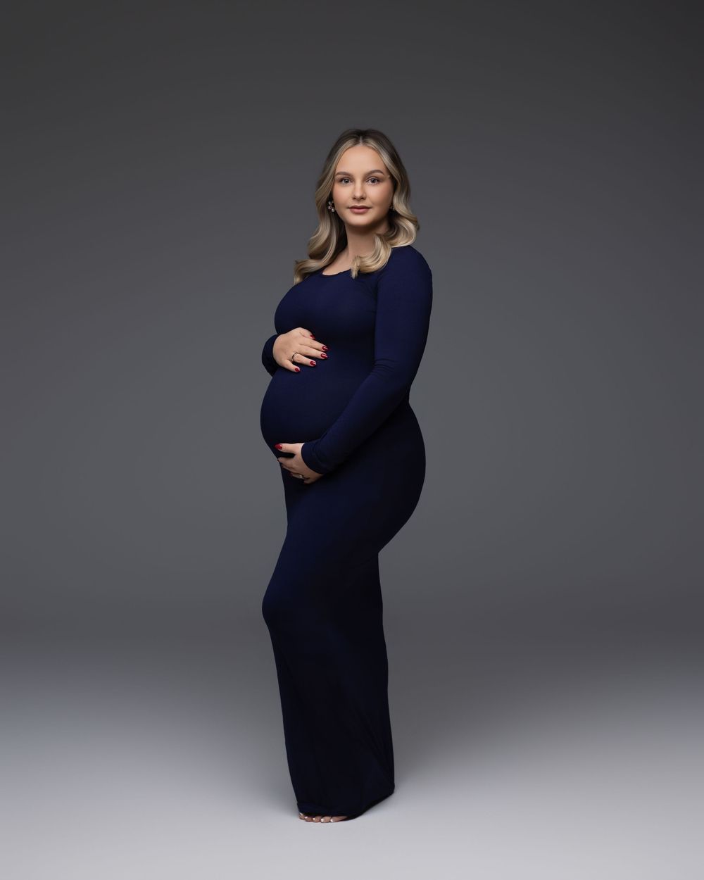 Expectant person in a fitted navy blue long dress poses elegantly against a dark gray studio backdrop.