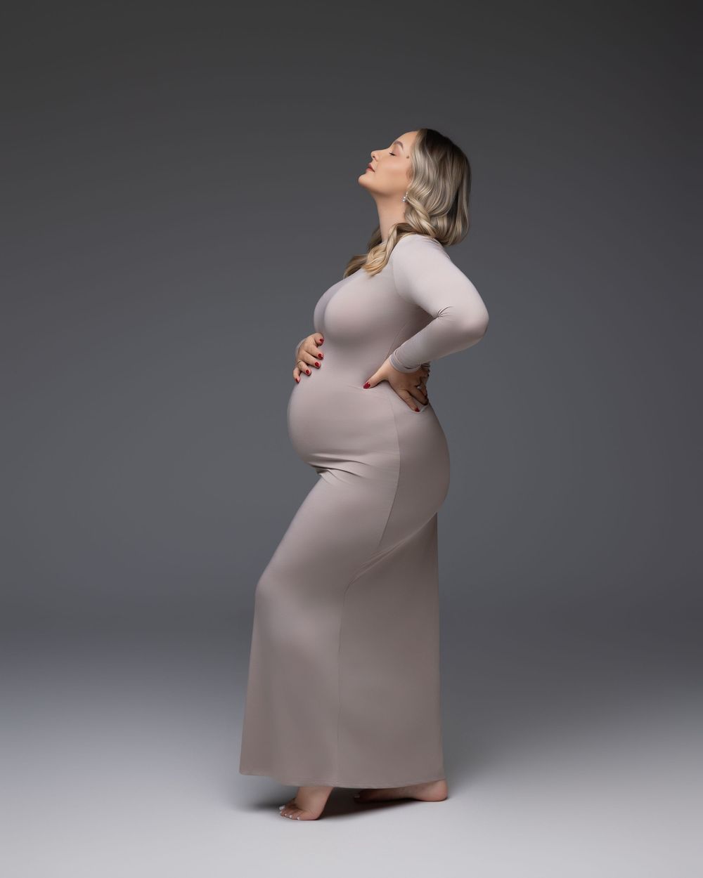A pregnant woman in a form-fitting beige dress poses in profile against a gray studio backdrop.