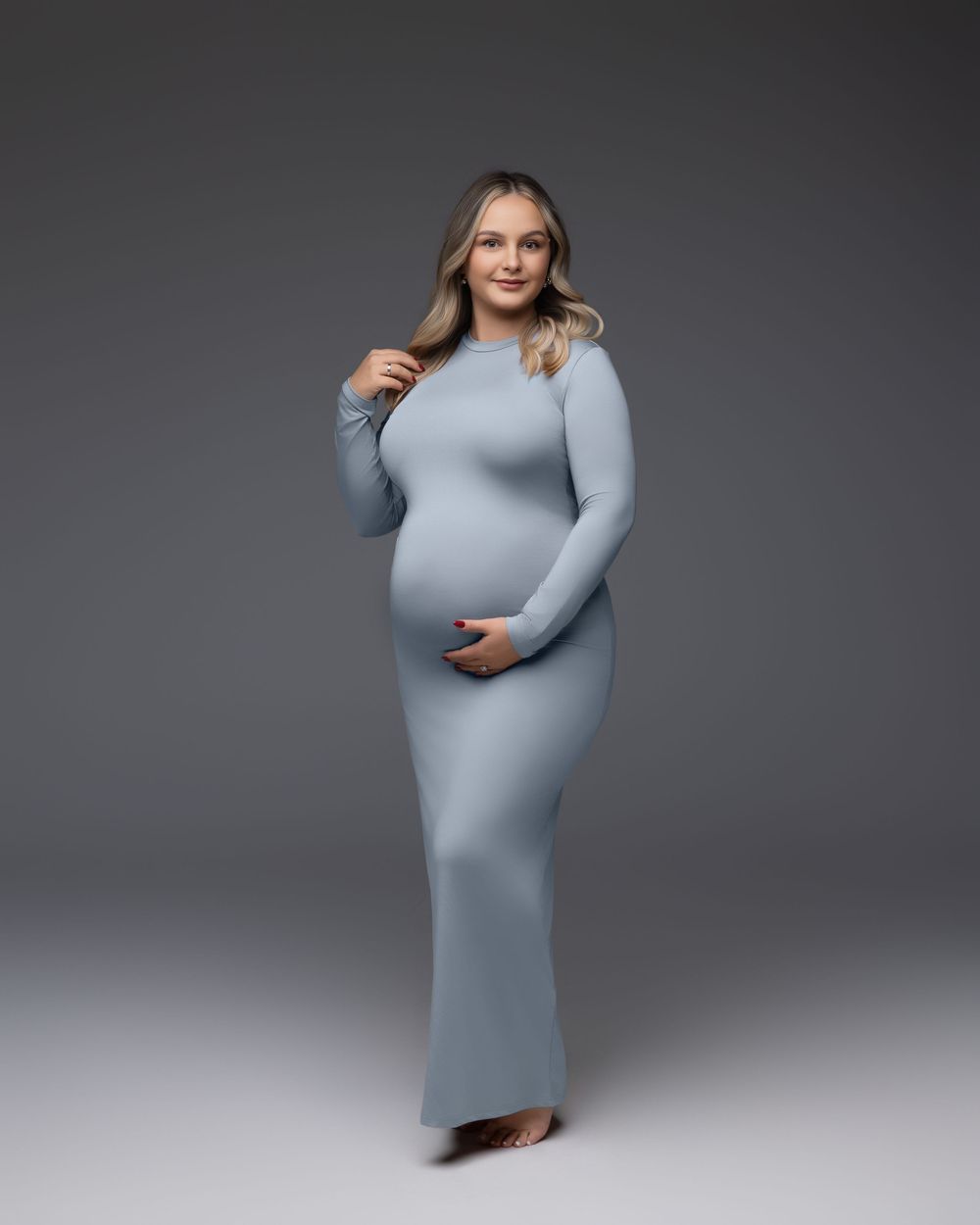 Side view of an expectant mother wearing a form-fitting light gray dress photographed against a gray studio backdrop.