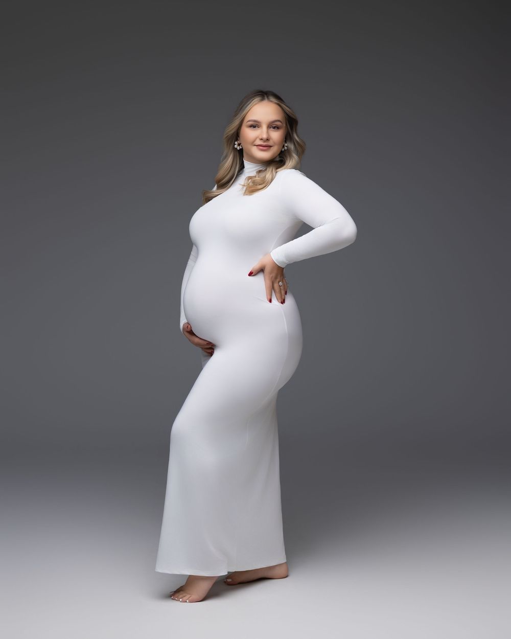 An expectant figure poses in a fitted white long sleeve maxi dress against a gray studio backdrop.