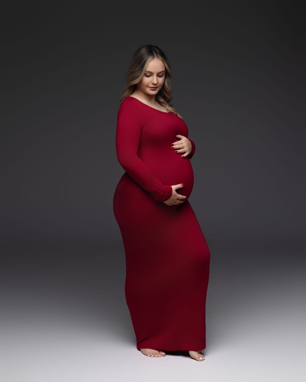 A person wearing a long burgundy maternity dress poses during a professional photo shoot against a grey backdrop.