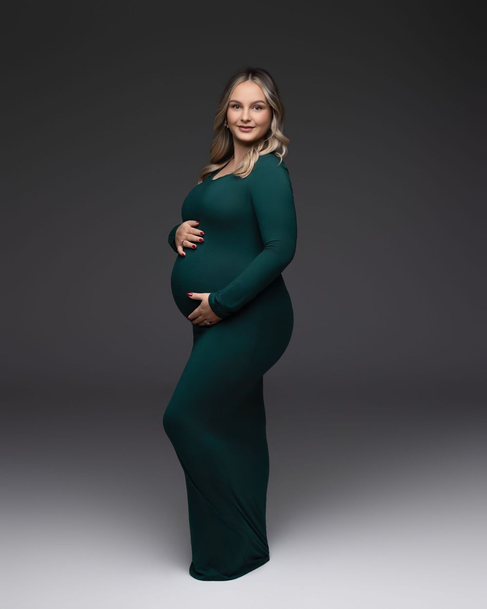A person in a fitted emerald green long sleeve dress poses against a dark gray backdrop during a maternity photoshoot.