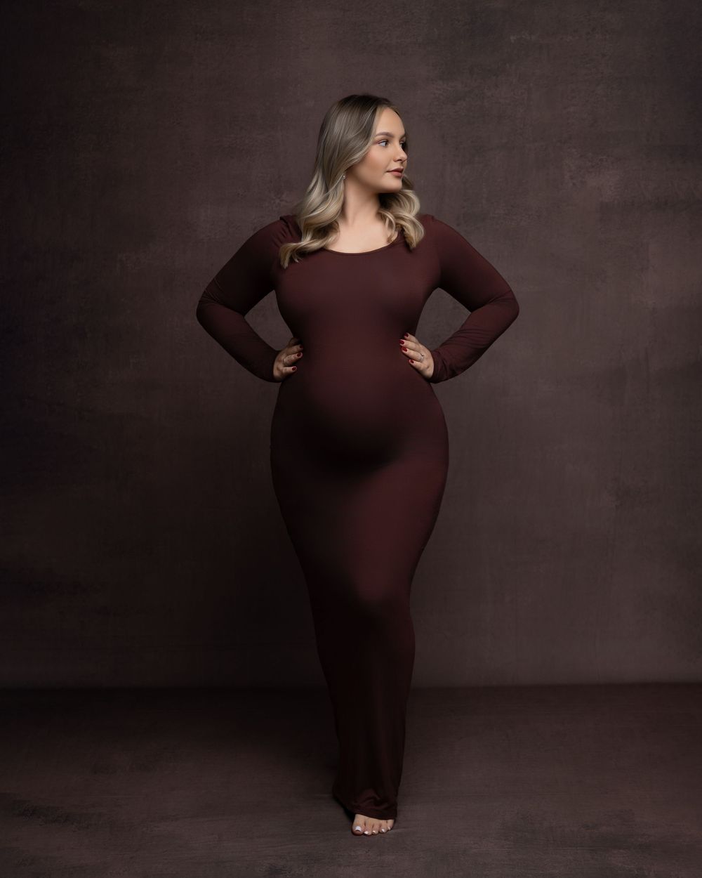 Dramatic maternity portrait series shows woman in brown dress against dark background posing with elegance.