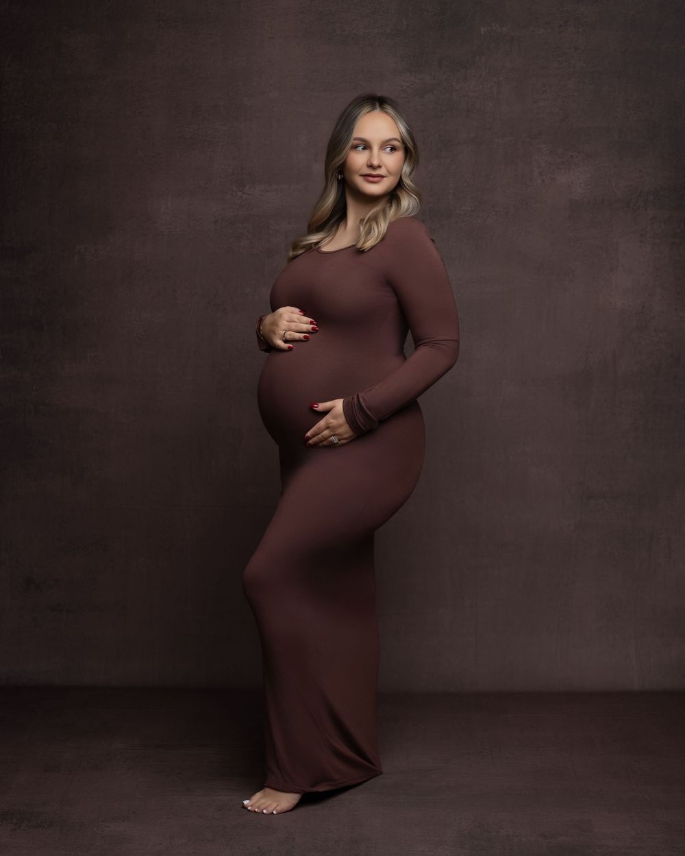 Artistic maternity photos showcase expectant mother in burgundy dress against moody studio backdrop.