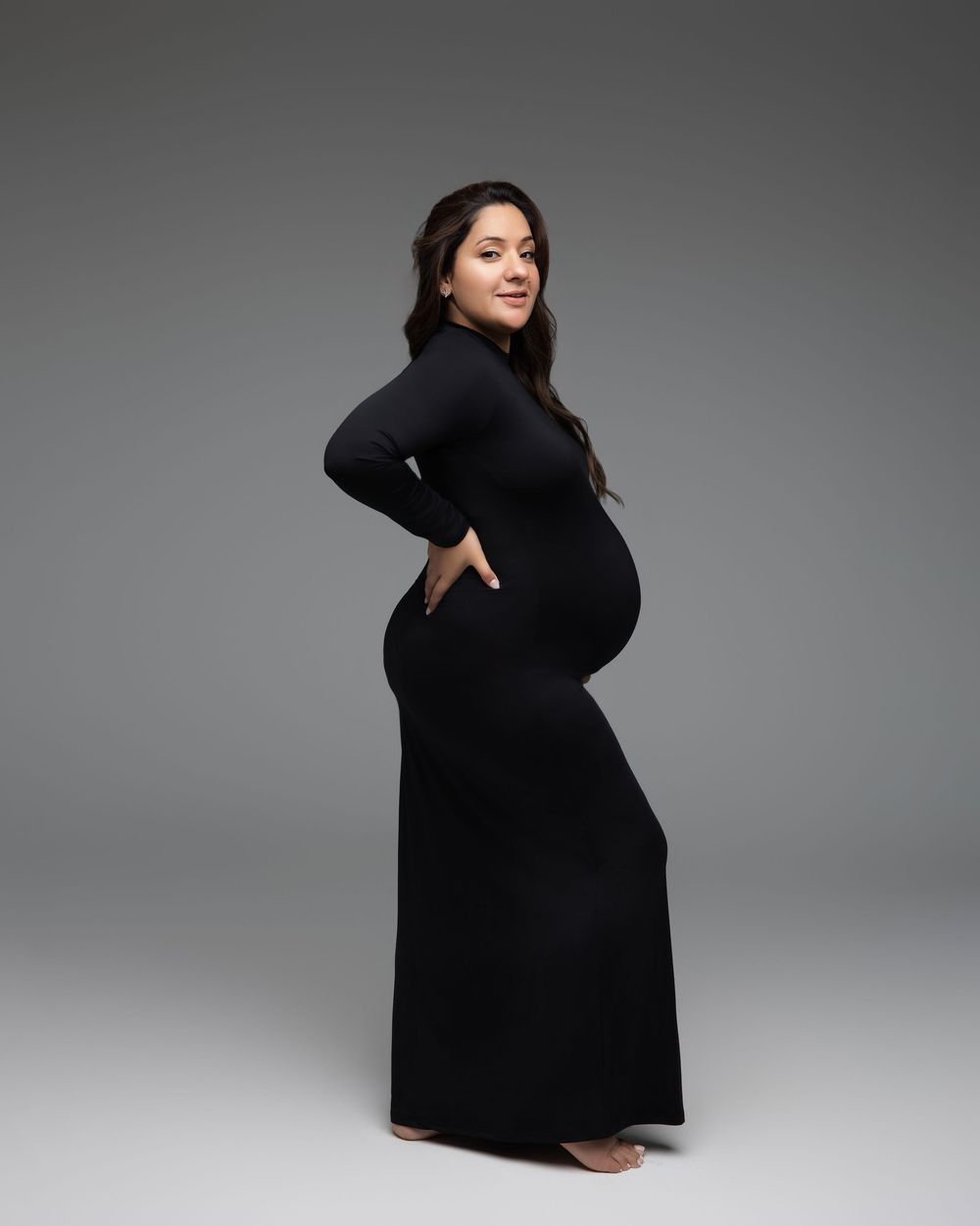 A pregnant figure in a long black dress poses elegantly against a gray studio backdrop.