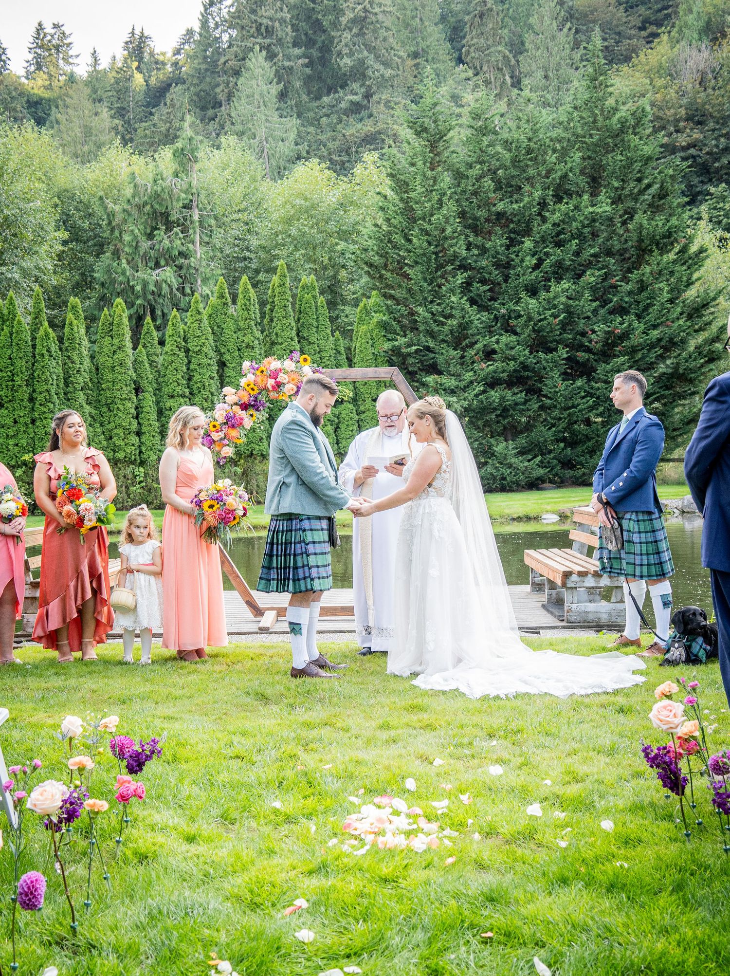 Scottish-themed outdoor wedding ceremony in a garden setting with colorful florals and evergreen backdrop.