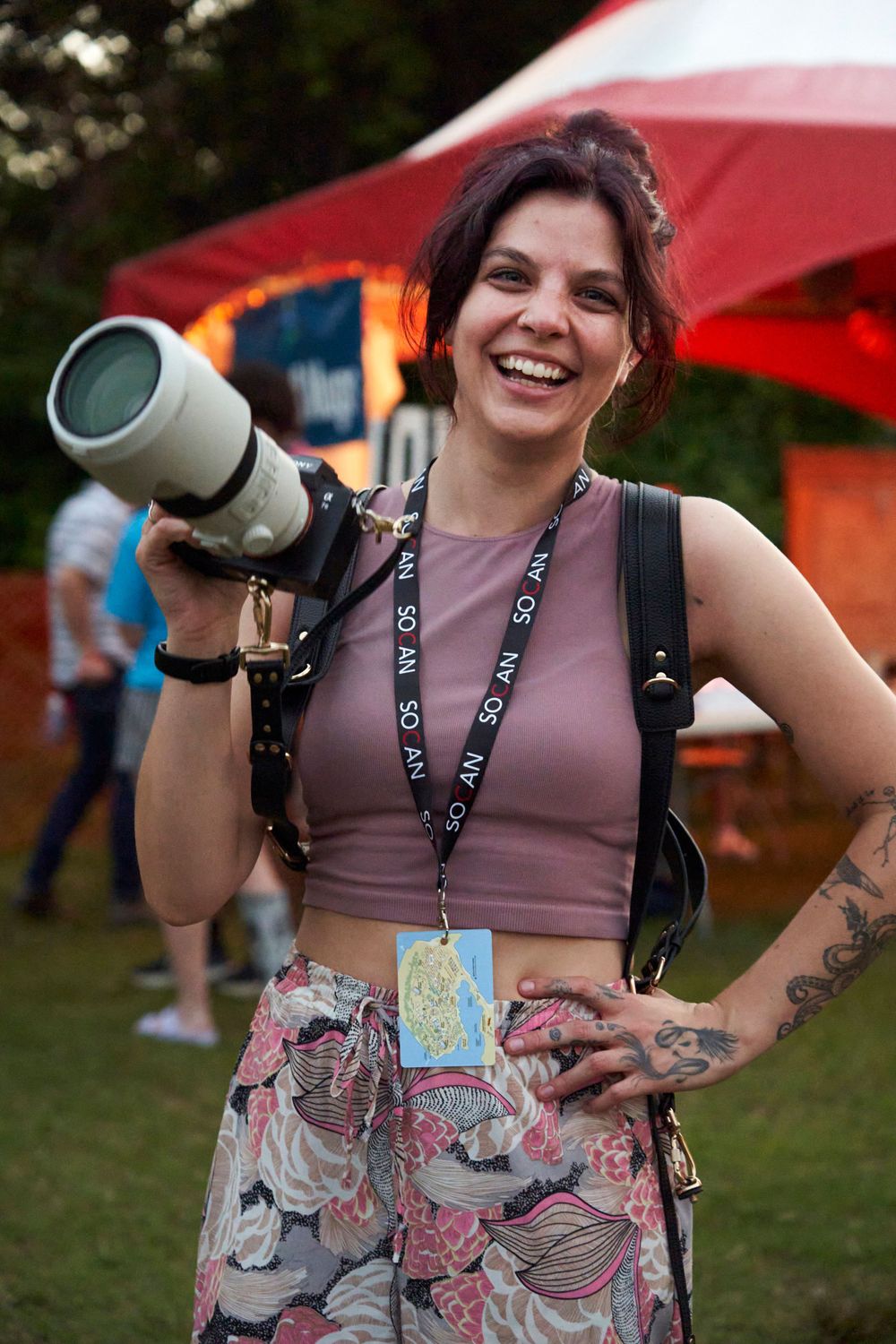 Professional photographer with camera gear and festival credentials smiles at outdoor event with red tent in background.