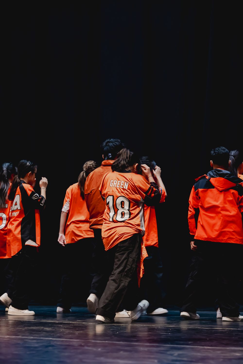 Dance group in matching orange jerseys performing on dark stage.