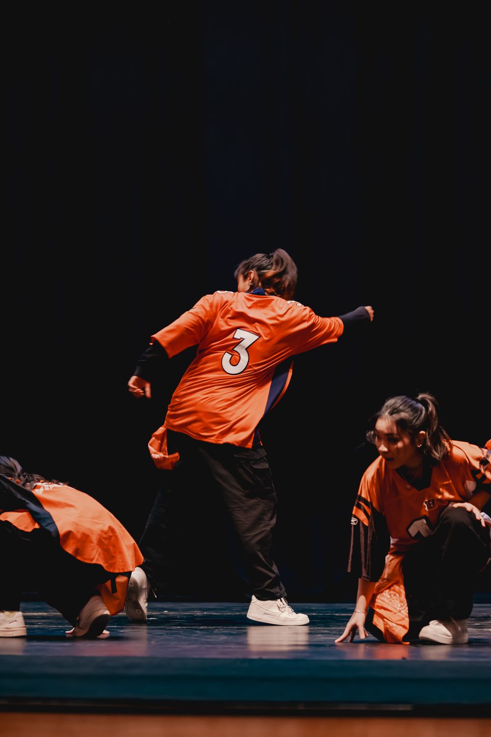 Dancers in bright orange jerseys perform dynamic hip hop routine on stage.