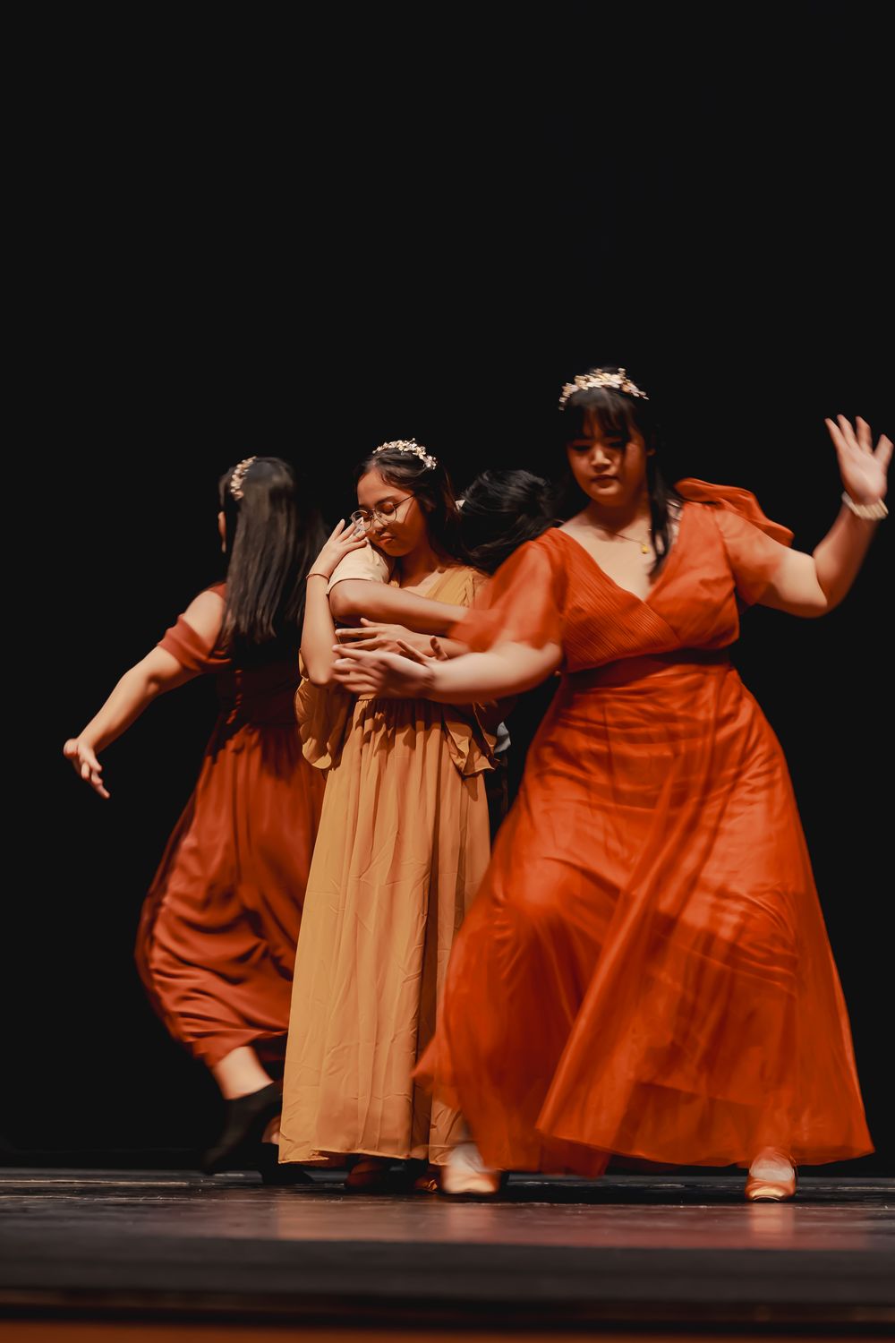 Dancers in flowing orange and beige dresses perform choreographed routine on stage.