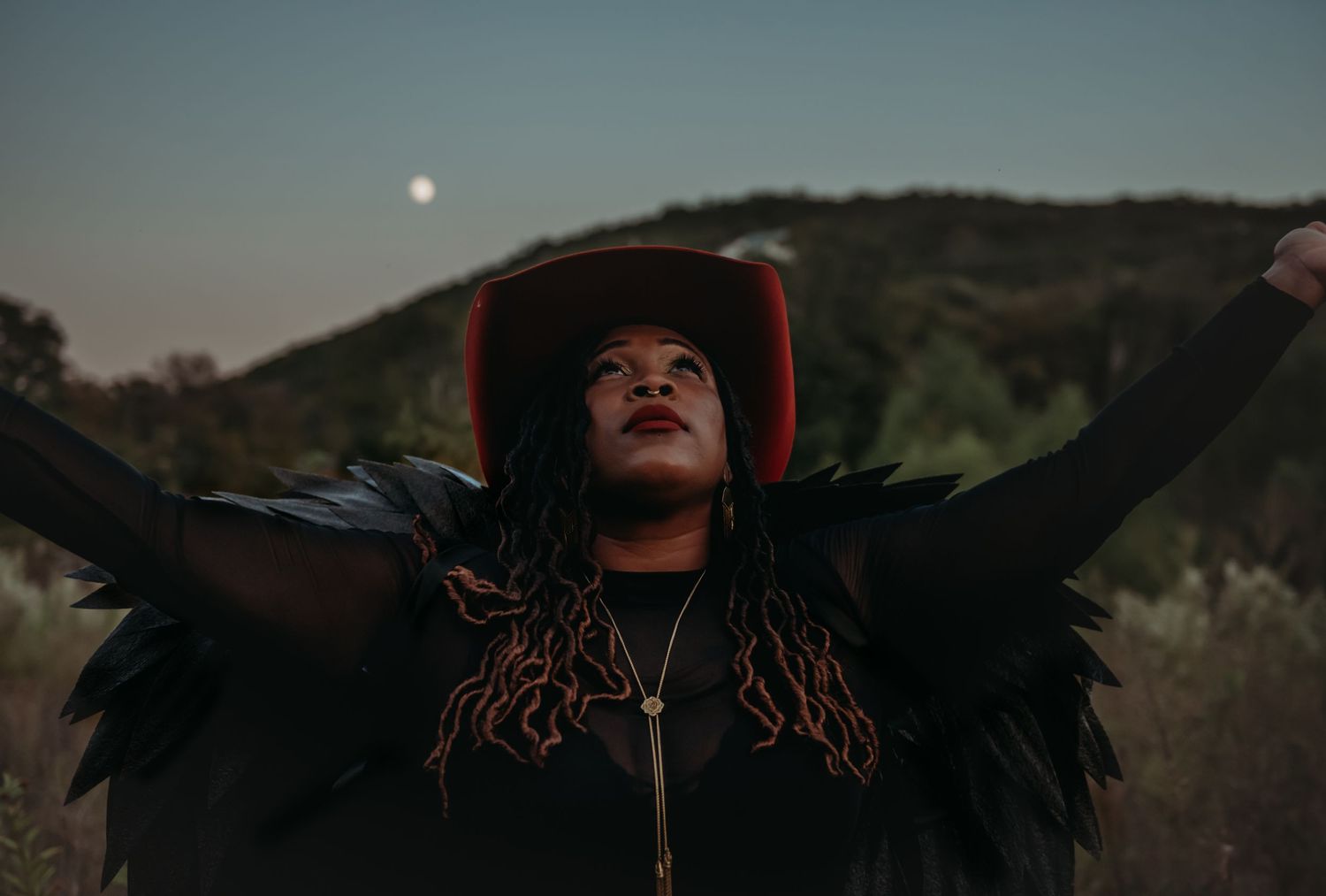 A person wearing a black feathered jacket and wide brimmed hat poses with arms outstretched under a moonlit sky.