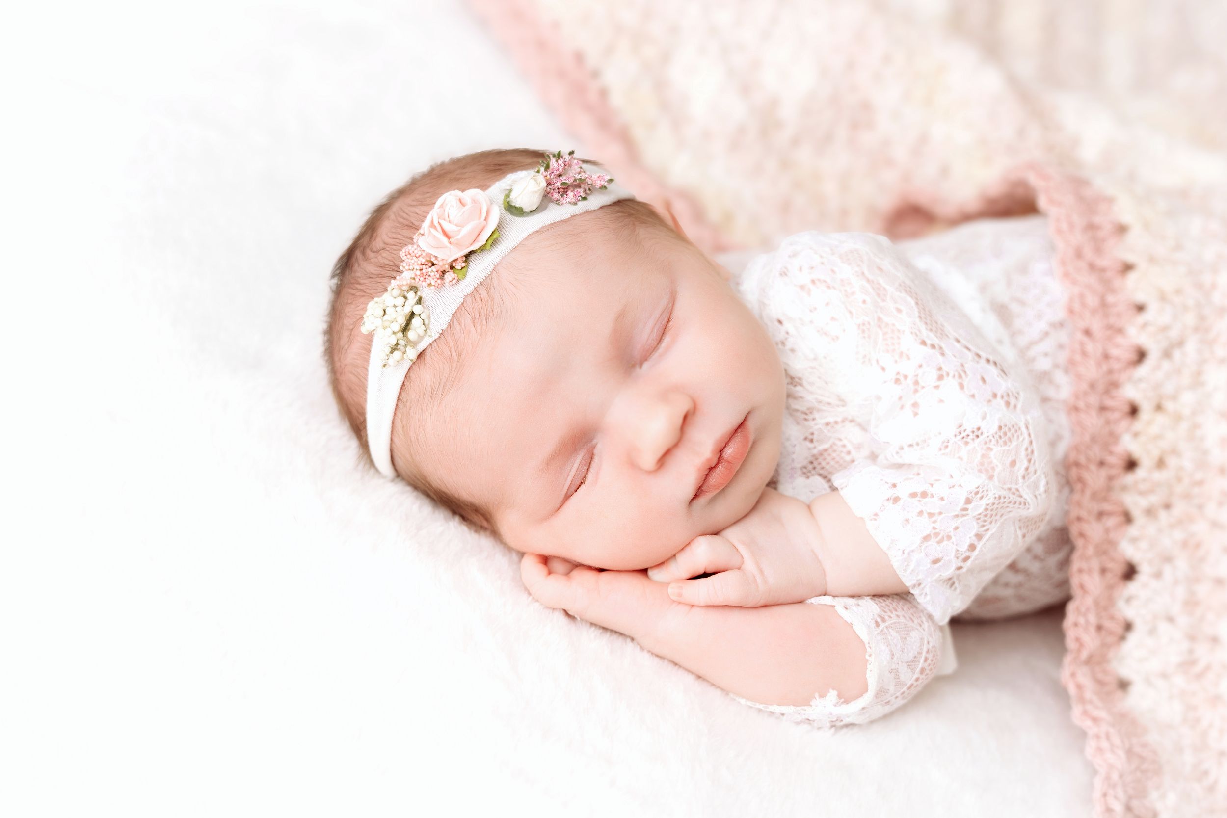 Newborn baby sleeping peacefully on soft white blanket wearing delicate lace headband.