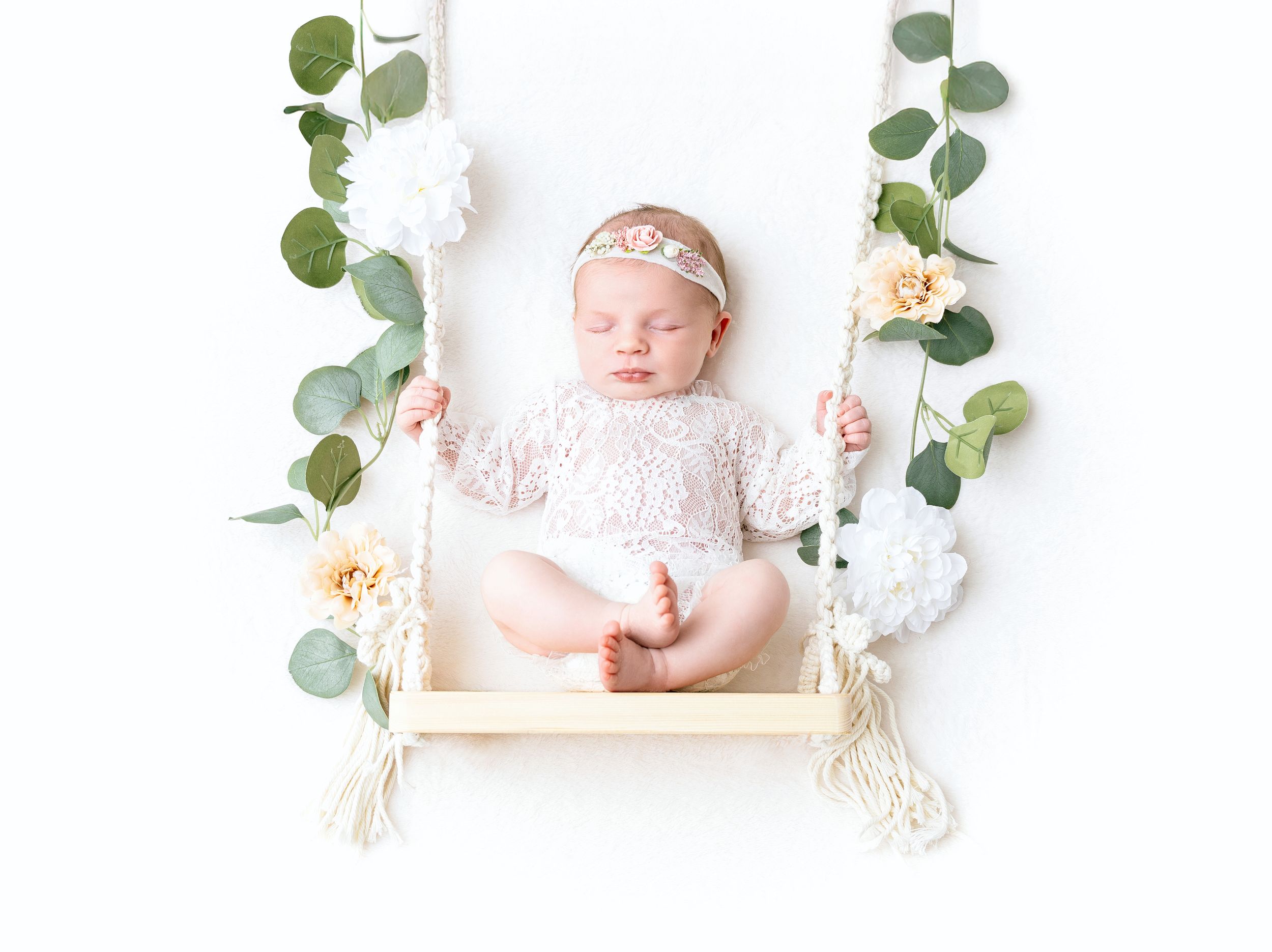 Sleeping baby on white wooden swing decorated with eucalyptus leaves and peach roses.