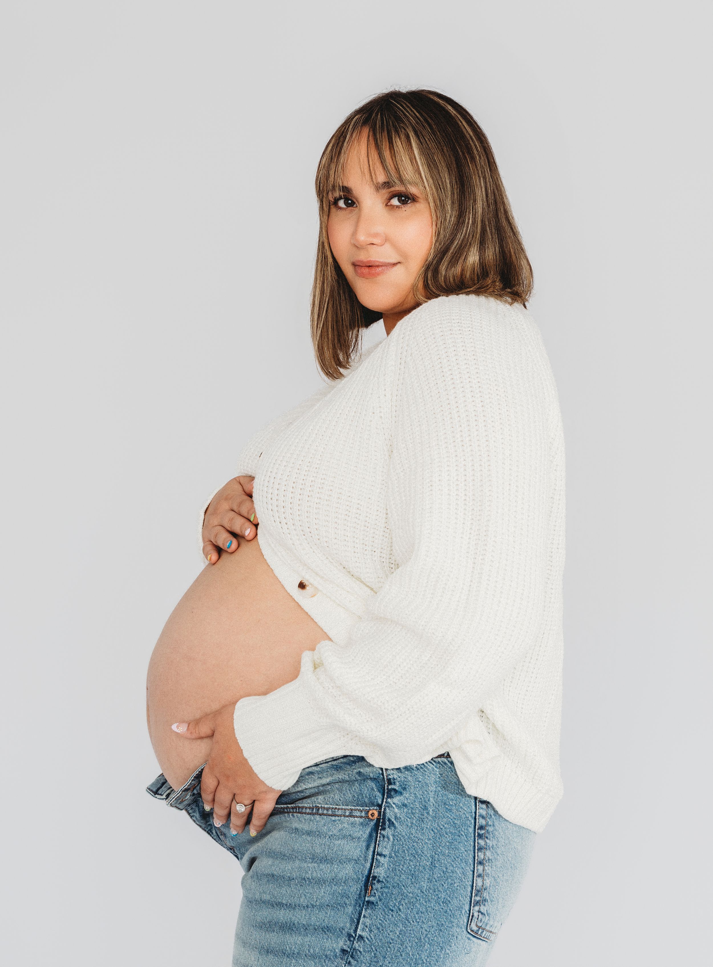 Pregnant woman in white knit sweater and jeans cradling her baby bump, posing sideways in a studio.