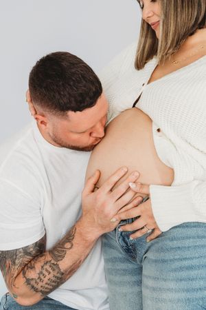 A tattooed man kisses a pregnant woman's bare baby bump during a maternity photo shoot.