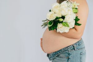 Pregnant woman in jeans holding white rose bouquet against light background, side profile maternity photo.