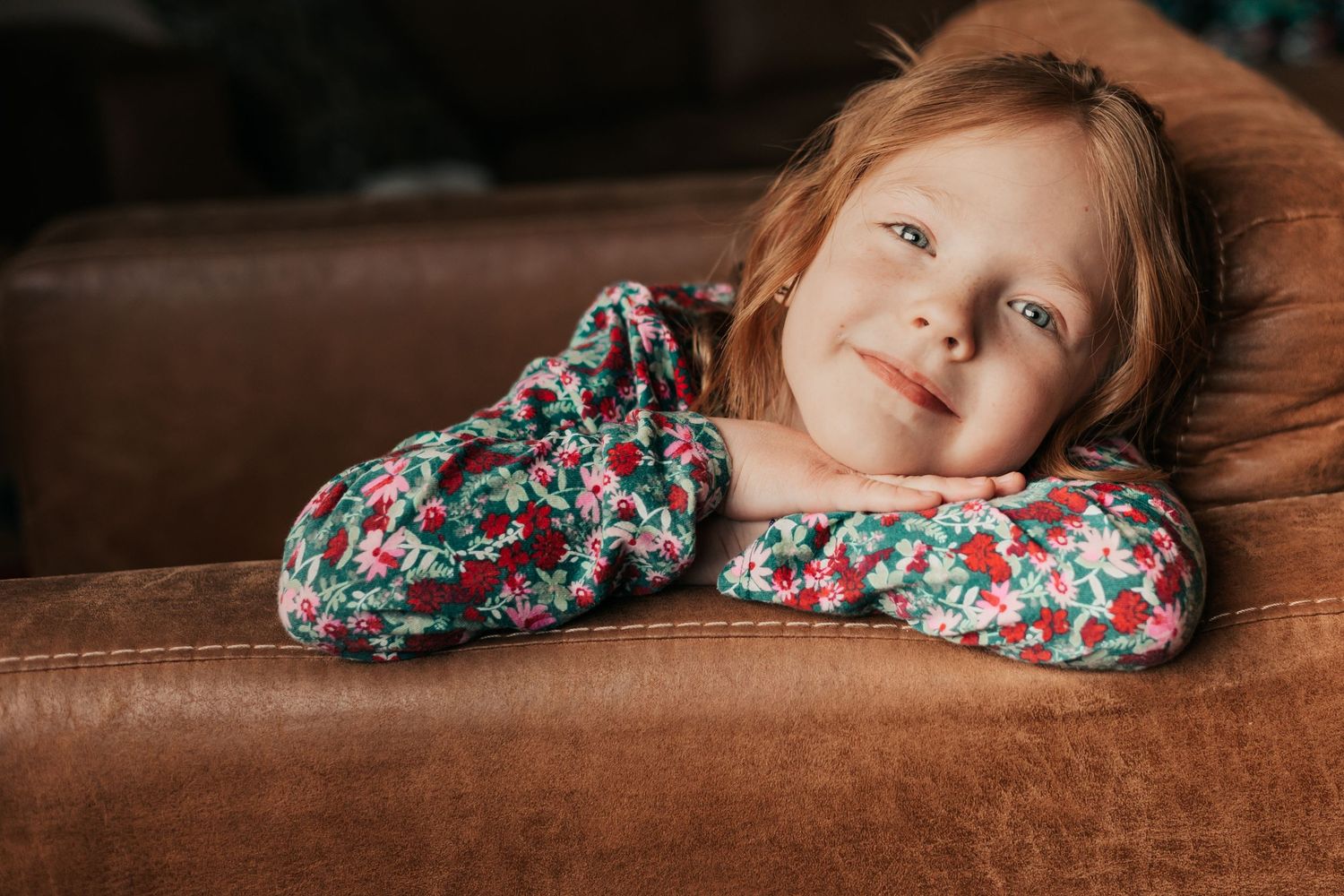 Little girl with red hair smiling and resting on a brown leather couch | In-Home Family Photos in Westminster, CO