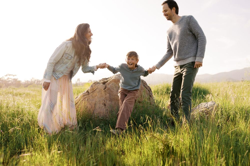 Family walking together through a sunlit meadow at golden hour wearing light colored clothing and holding hands.