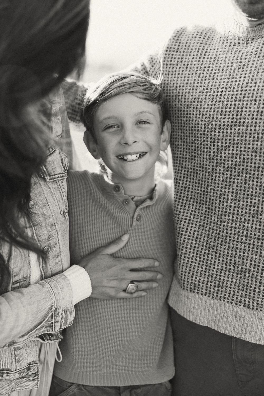 Black and white portrait of a young child smiling while standing between two adults in casual clothing.