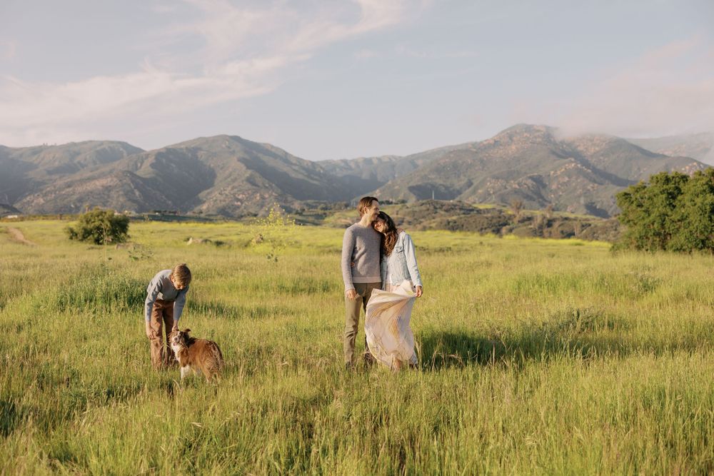People walk with their dog across a scenic mountain meadow with rolling hills and green grass in the background.