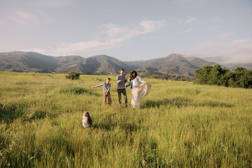 A panoramic view of rolling hills and mountains with people walking through a grassy meadow at sunset.