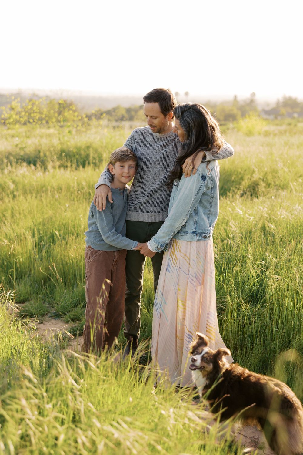 Family enjoying a tender moment together in a sunlit meadow with their Australian Shepherd dog.