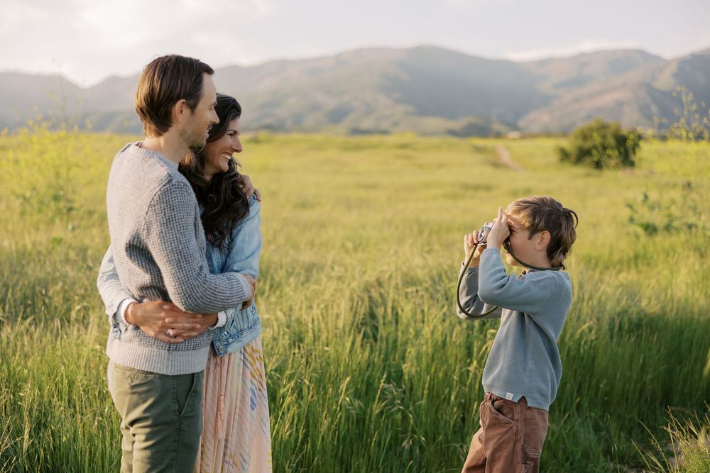 Family enjoying a tender moment together in a sunlit meadow with rolling hills in the background.