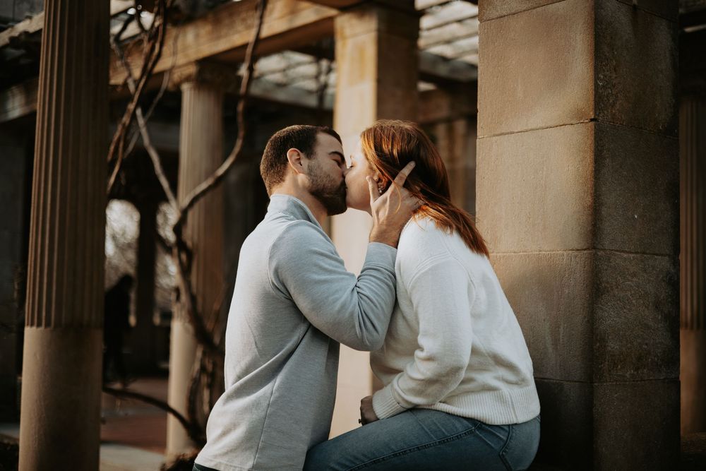 A couple shares an intimate moment while sitting together against a stone pillar in warm lighting.