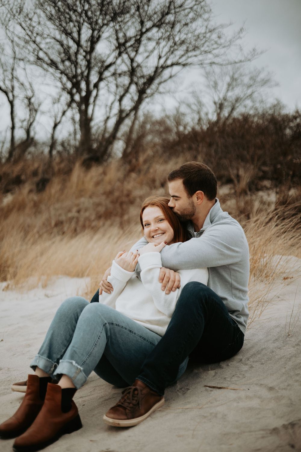A couple shares an intimate moment sitting together on sandy ground surrounded by tall grass and bare trees on an overcast day.