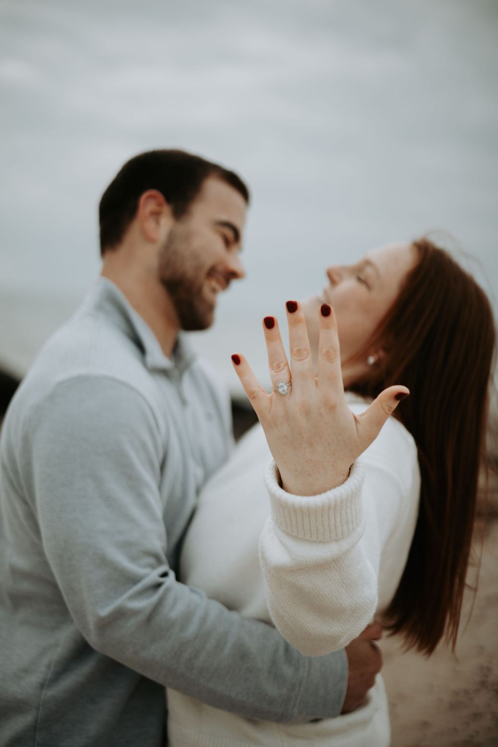 A couple embraces on a cloudy day as she displays her engagement ring with burgundy nails.