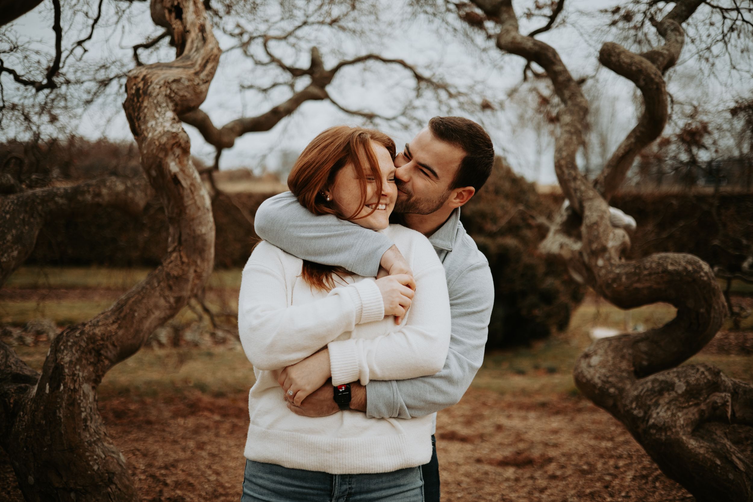 Couple in white sweaters sharing intimate moments during outdoor autumn photoshoot among rustic trees.