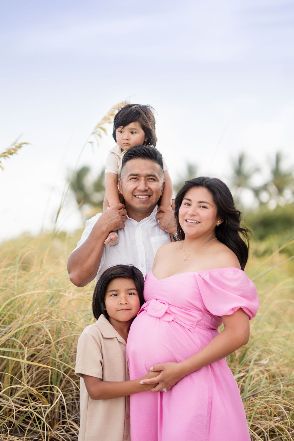 Happy Hispanic family with pregnant mother in pink dress poses outdoors in golden sea grass field.