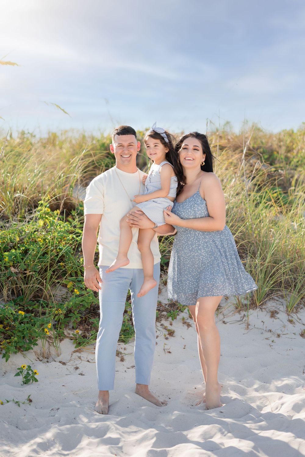 Smiling family of three standing on white sand beach with sea oats and blue sky behind them.