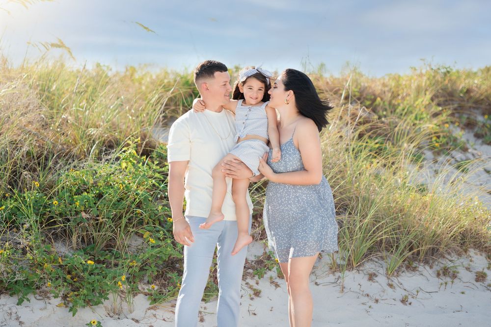 Happy family of three posing together on sandy beach dunes with tall grass in background.