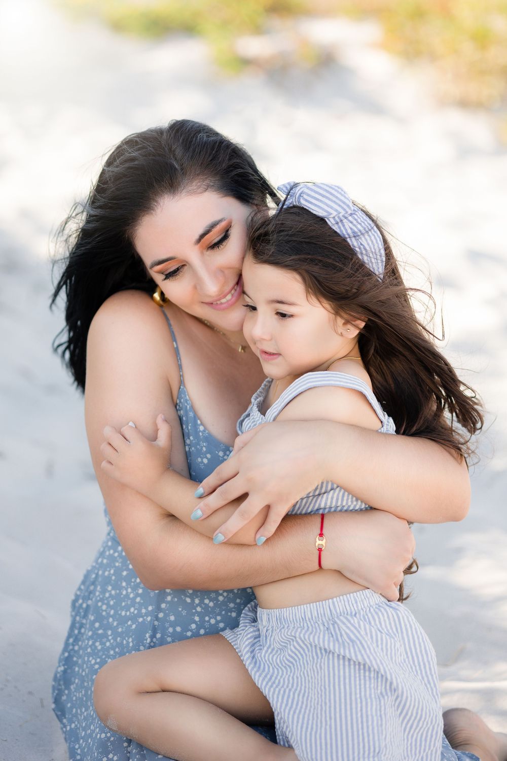 Mother and daughter sharing tender embrace while sitting on sandy beach in matching blue outfits.